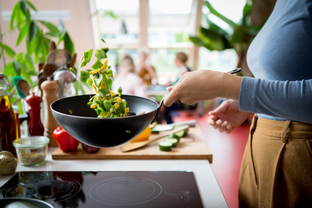 Een afbeelding waarop een dame in de keuken staat en de groente in de wok schud